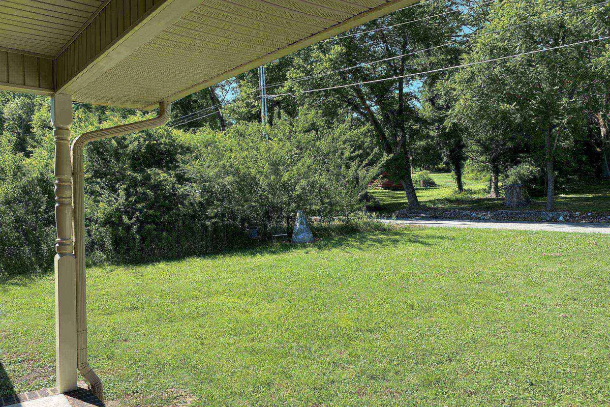 A welcoming front porch framed by sturdy posts, with lush greenery and an open lawn leading to a dirt path. The scene exudes tranquility, inviting guests to relax on the porch or stroll through the peaceful neighborhood.