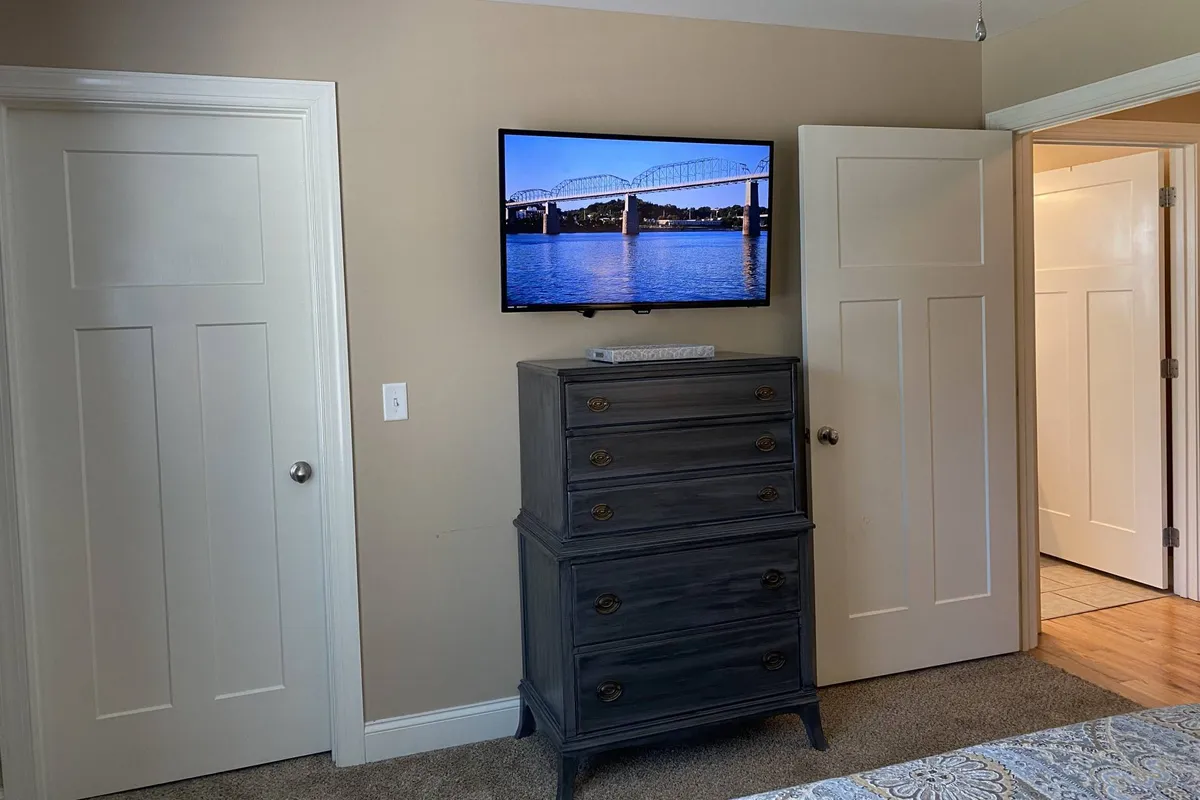 A cozy interior scene with a TV displaying a picturesque bridge over water, set above a dressed dresser. The mood is peaceful and inviting, ideal for relaxing or unwinding after a day out.