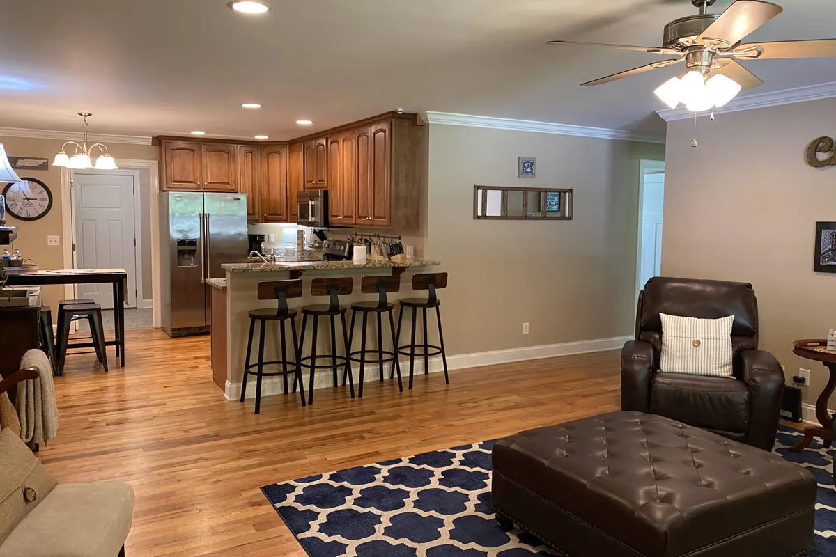 A view of a spacious kitchen island with bar stools and a plush recliner in the background. The area is well-lit, featuring inviting light fixtures and a cozy rug, creating a seamless flow for entertaining or relaxing.