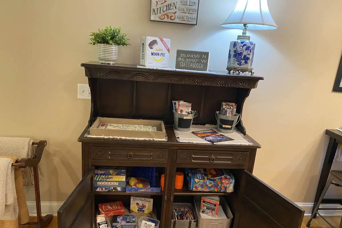A wooden writing desk surrounded by kitchen-themed decor, a decorative lamp, maps, brochures, and a visible selection of games and toys, all situated in a warm, cozy corner. The scene evokes family gatherings, game nights, and planning adventures.
