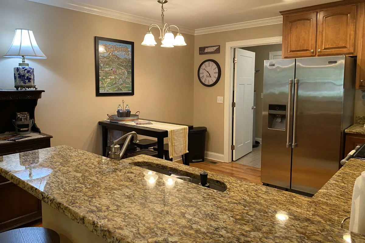 A cozy kitchen and dining area, complete with a central island featuring granite countertops, a chandelier above, modern appliances, and wall artwork. The scene is bright and welcoming, emphasizing the space's dual purpose for both meal preparation and communal dining.