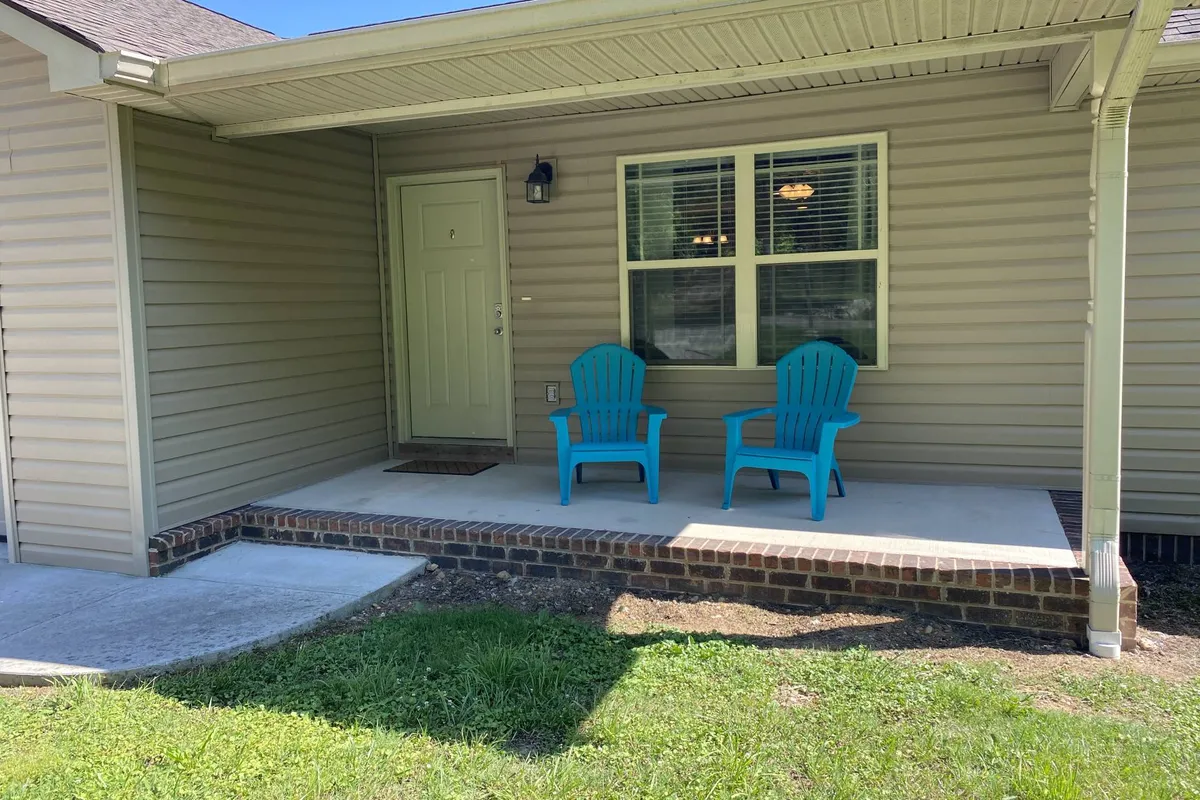 The front exterior of the house with two inviting turquoise chairs positioned on a small porch, next to a light green door and a window with blinds. The setting suggests a relaxed, welcoming entry point for family gatherings or quiet moments after outdoor adventures.