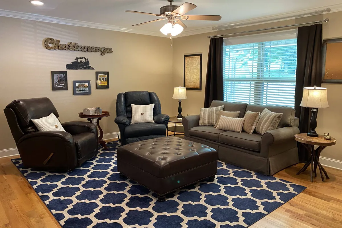 The image showcases a warm and inviting living room with plush leather recliners, a cozy sofa adorned with decorative pillows, a stylish coffee table, and a soft large area rug. The seating is arranged to encourage conversation or relaxation, perfect for gatherings, movie nights, or unwinding with a book by the window.