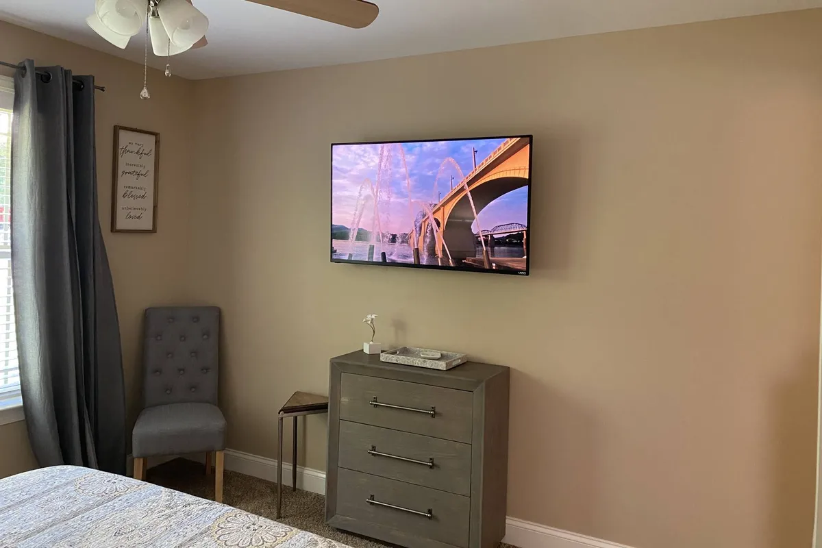 A cozy corner of a bedroom or guest room, featuring a wall-mounted TV displaying a scenic sunset, a soft gray dresser, and a comfortable chair. The space is designed for leisure, perfect for watching TV or unwinding after a busy day.
