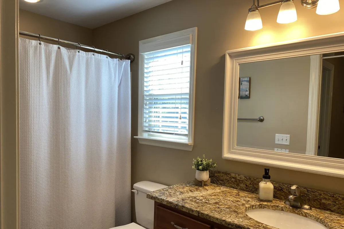 This image features a calm and refreshing bathroom interior with a granite countertop, a large mirror, a shower curtain, and a window allowing in peaceful morning light. The space exudes a clean, well-maintained atmosphere ideal for daily routines and self-care moments.