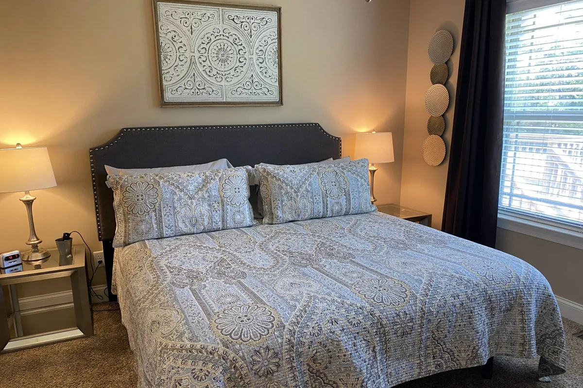 This image showcases a bedroom with a comfortable bed featuring decorative bedding, wall art above the headboard, and soft table lamps on either side. Gentle sunlight filters through the blinds, creating a warm and inviting atmosphere ideal for relaxation.
