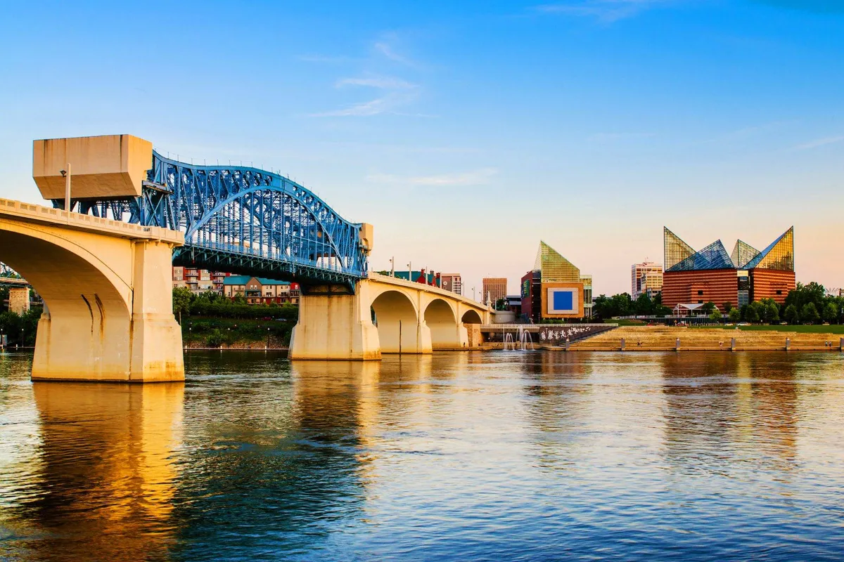 A tranquil river scene featuring a distinctive blue arch bridge, modern downtown Chattanooga buildings in the background, and calm waters reflecting the cityscape. The setting sun adds a warm glow, hinting at a vibrant and scenic urban environment.